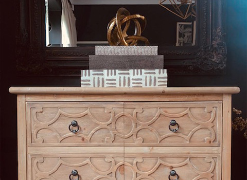 A wooden dresser with ornate carvings, featuring two drawers and decorative knobs, topped with a stack of boxes and a golden sculpture, reflecting a fashionable home decor style.